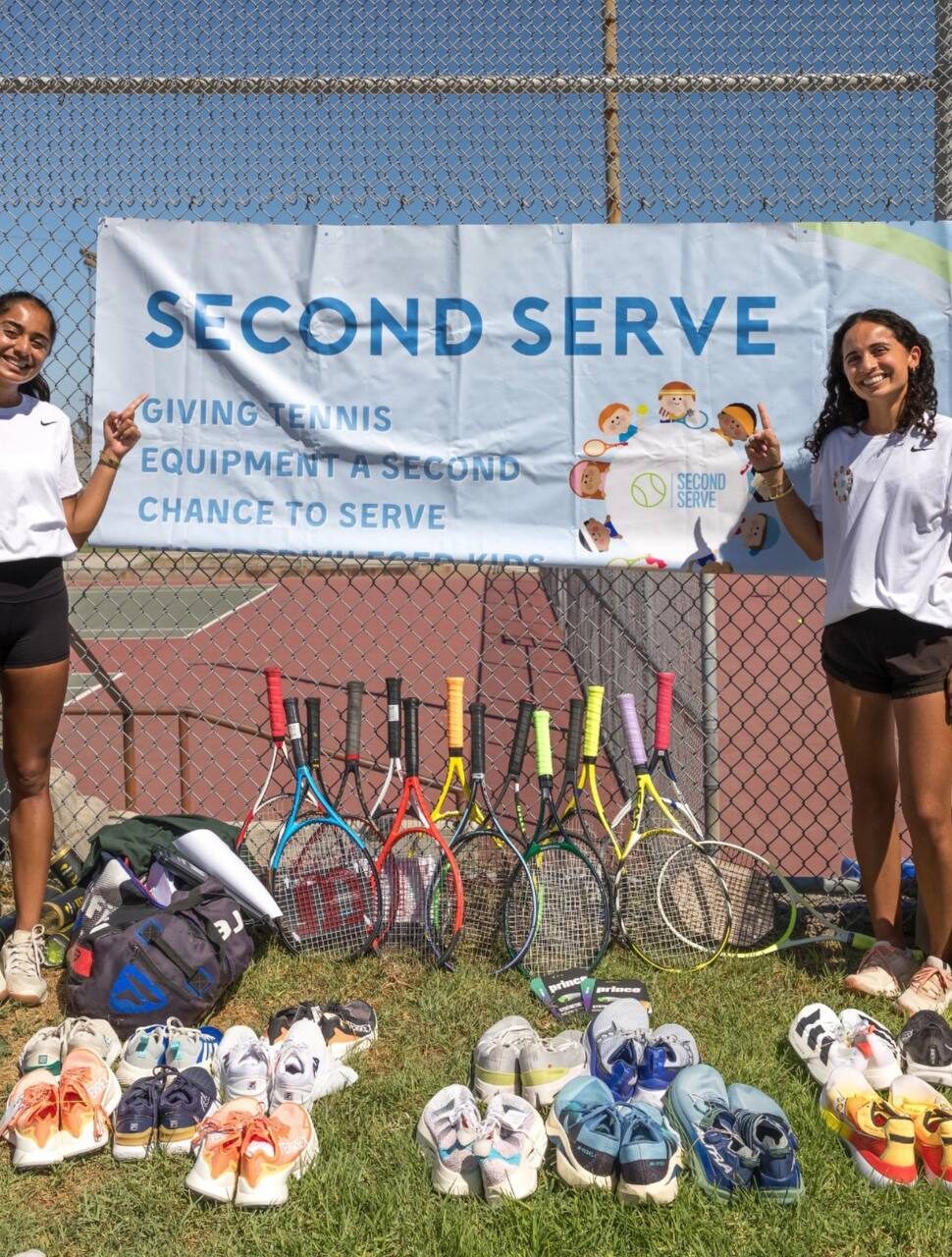 Two Second Serve volunteers beside a banner at a public tennis court, with donated rackets and shoes lined up on the grass