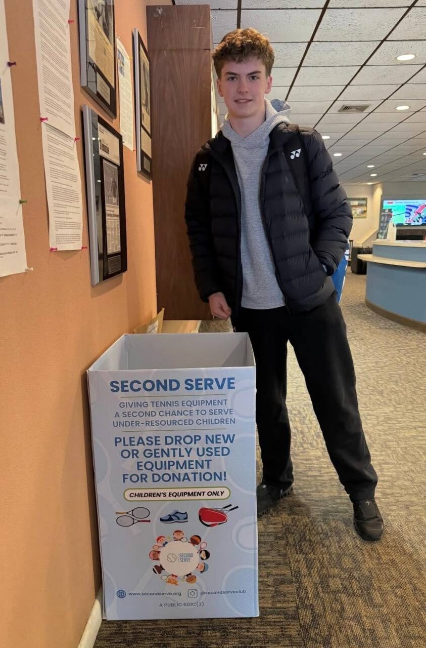 A teenage player standing next to a Second Serve collection box in a tennis club lobby